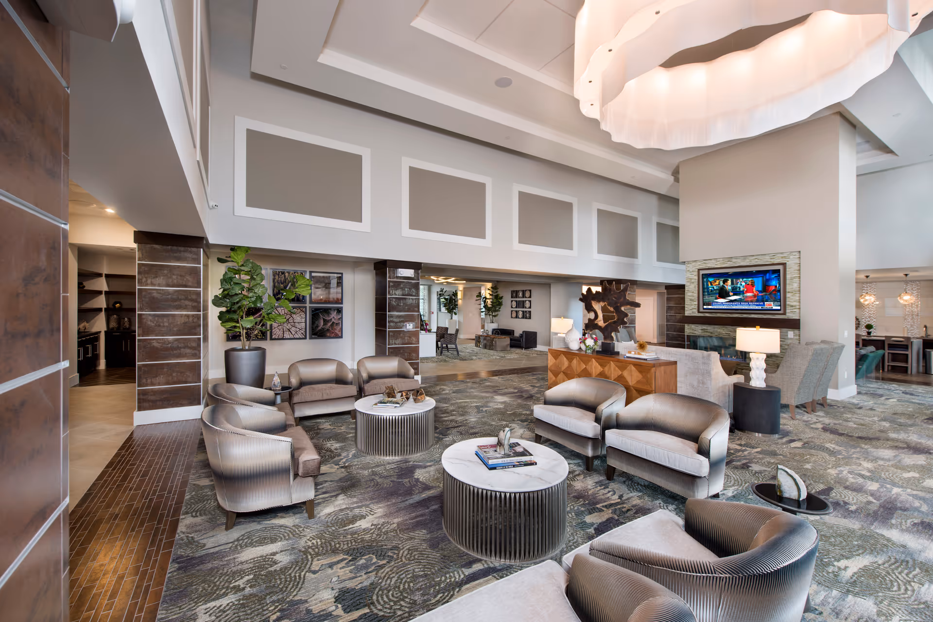A spacious and modern living room area with multiple seating arrangements including armchairs and loveseats around circular coffee tables. The room features a large decorative ceiling light, a flat-screen TV mounted above a fireplace, and decorative art pieces on the walls. The carpet has a patterned design, and there are plants and lamps adding to the cozy ambiance.