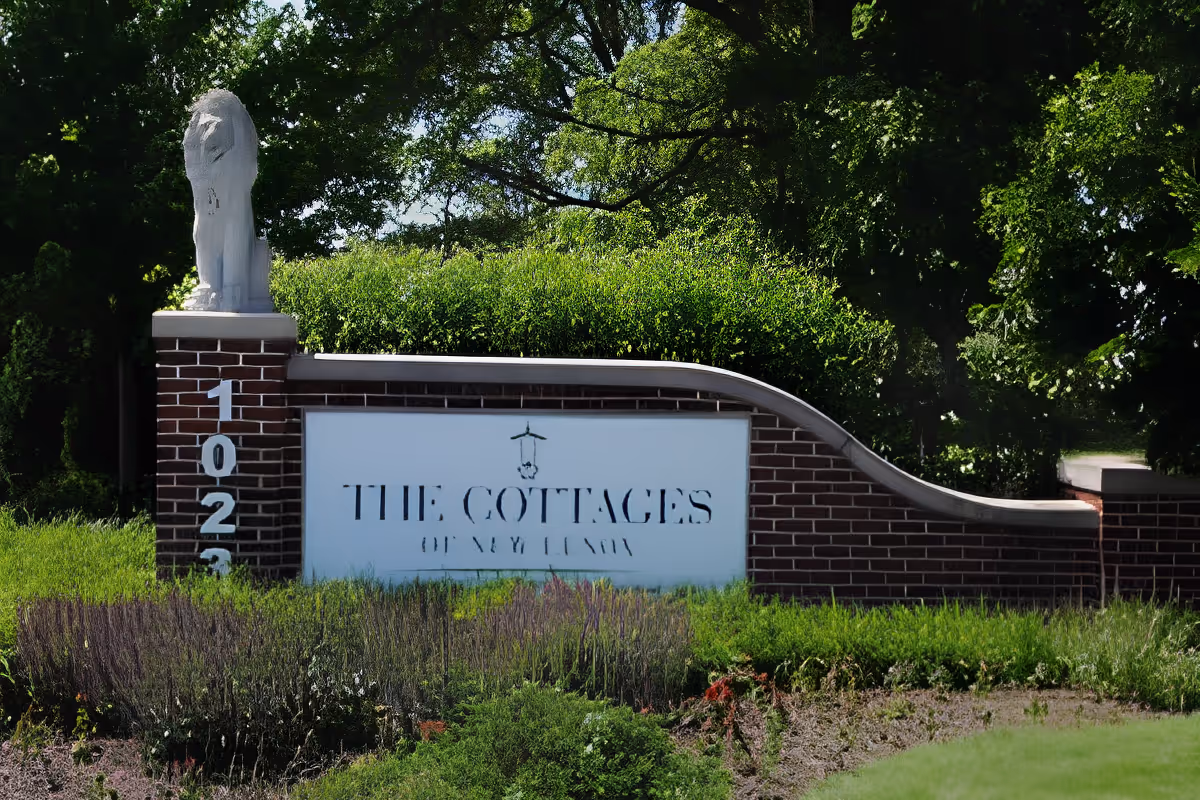 Brick entrance sign for The Cottages of New Lenox with address numbers on a pillar, a small lion statue, and surrounding greenery.