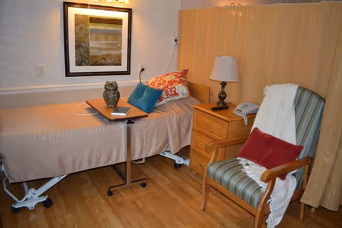 A senior living facility bedroom with a single bed covered in a beige bedspread, decorated with colorful pillows. Next to the bed is a wooden nightstand with a lamp and a telephone. A small table on wheels is positioned over the bed, holding a decorative owl and a tablet. A striped armchair with a red pillow and a white throw blanket is placed nearby. The room has wooden flooring and a framed picture on the wall.