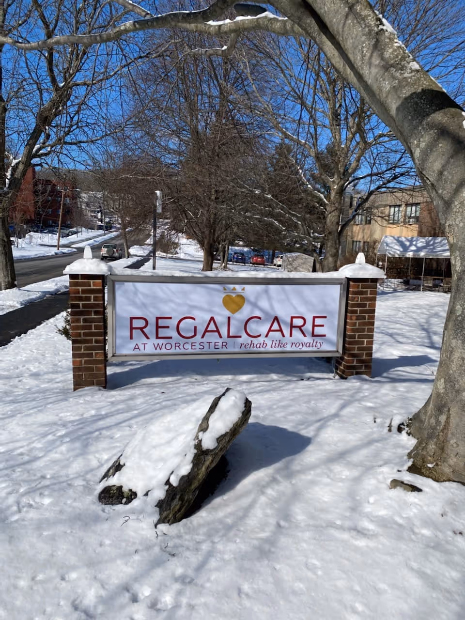 Outdoor scene showing a snow-covered ground with a sign for RegalCare at Worcester, which reads 'rehab like royalty'. The sign is supported by two brick pillars and there are leafless trees and buildings in the background under a clear blue sky.