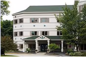 Front view of a three-story white building with a green roof and covered entrance surrounded by trees and shrubs.