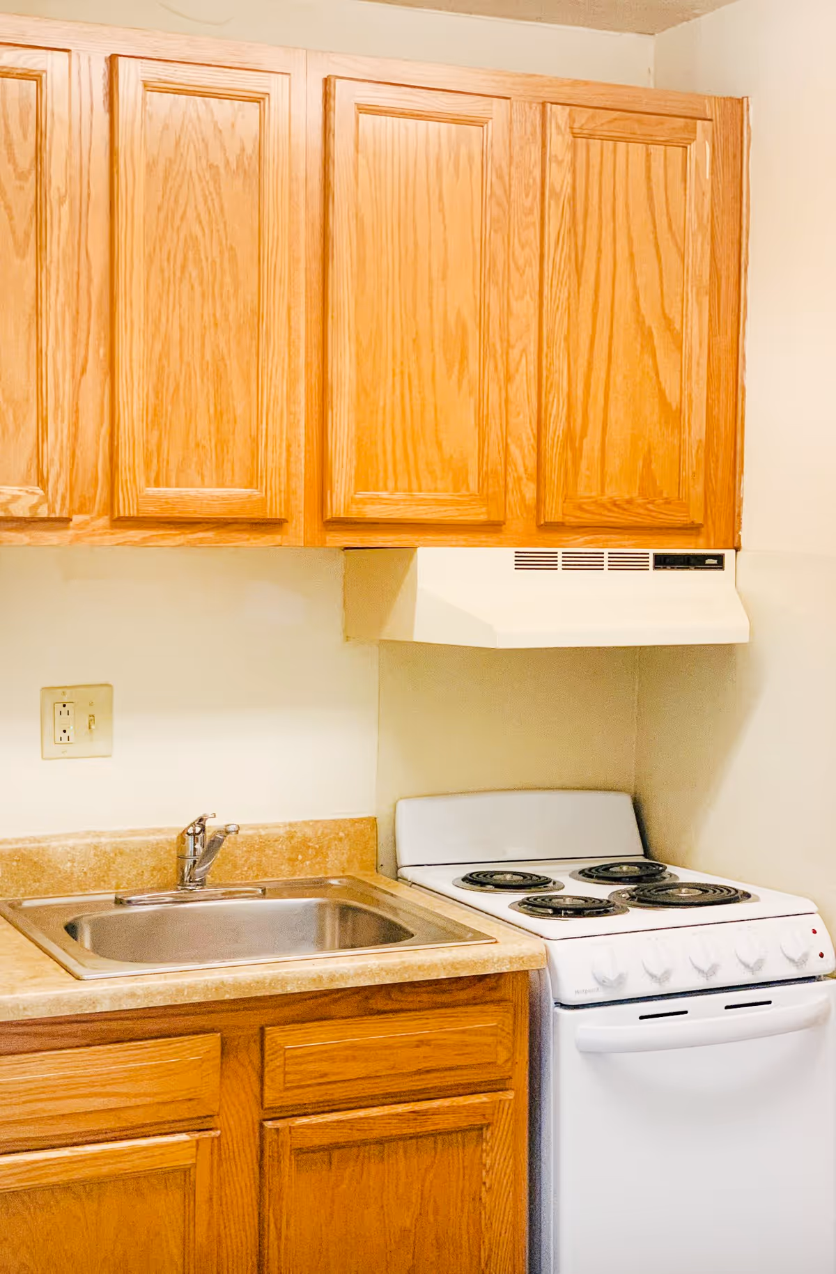 A small kitchen area with wooden cabinets, a stainless steel sink, a beige countertop, and a white electric stove with four coil burners and an overhead range hood.