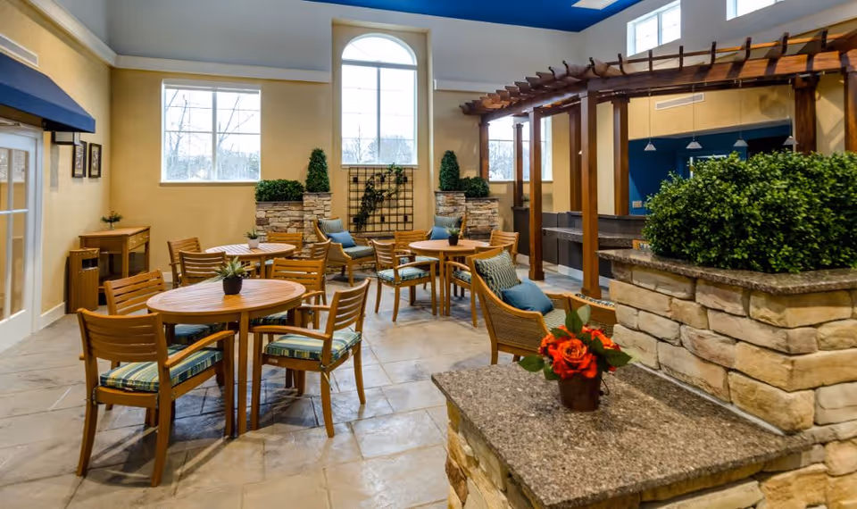 A bright and spacious indoor seating area with several round wooden tables and chairs with striped cushions. The room features large windows, stone planters with green plants, and a wooden pergola structure. There are decorative elements like small potted plants on the tables and a flower arrangement on a stone countertop in the foreground.