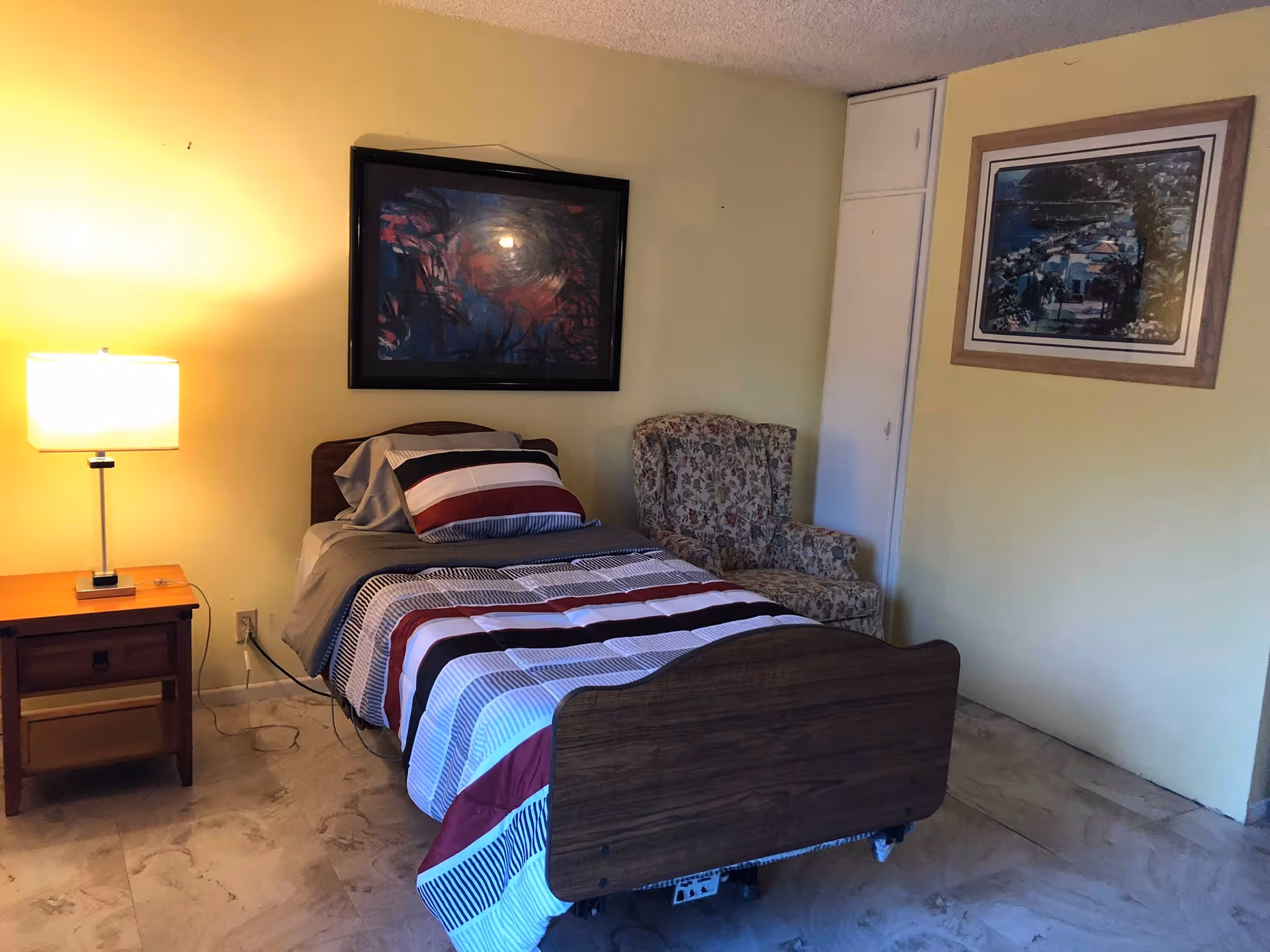 A small bedroom with a single bed covered in a striped comforter in shades of red, black, white, and gray. Next to the bed is a wooden nightstand with a lamp. There is a floral upholstered armchair in the corner, and two framed pictures hang on the yellow walls. The floor is tiled with a light-colored marble pattern.