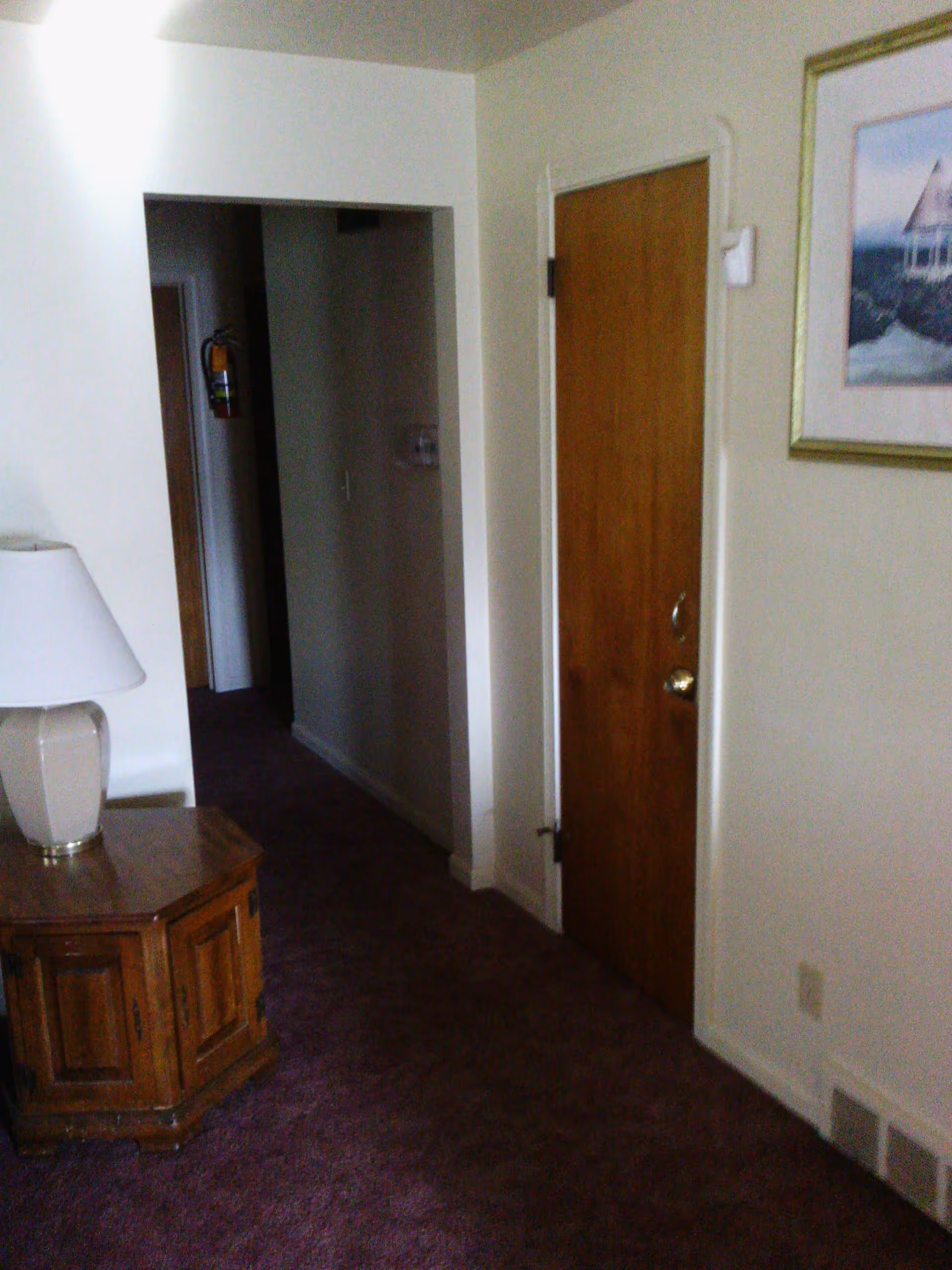 Interior hallway of a facility with a wooden door on the right, a small wooden side table with a lamp on the left, beige walls, purple carpet, and a framed picture hanging on the wall.