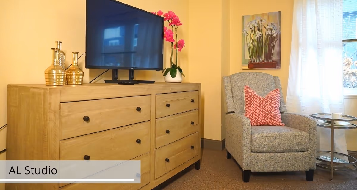 A cozy corner of a senior living facility room featuring a wooden dresser with six drawers, a flat-screen TV on top, two decorative gold vases, and a pink orchid plant. Next to the dresser is a comfortable gray armchair with a red patterned pillow. A small round side table with a cup and saucer sits beside the chair. The room has light yellow walls, a window with white curtains, and a floral painting hanging on the wall.
