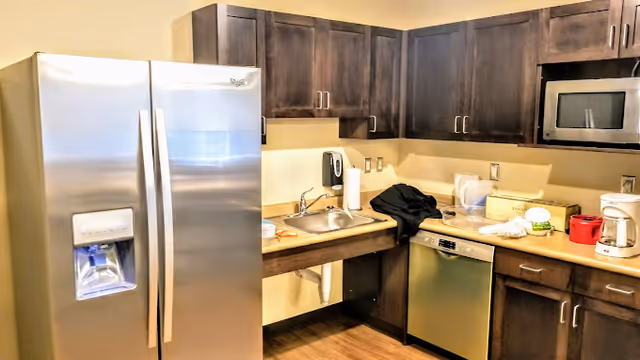 Compact kitchen with a stainless steel refrigerator, sink, dishwasher and microwave surrounded by dark wood cabinets and countertop appliances.