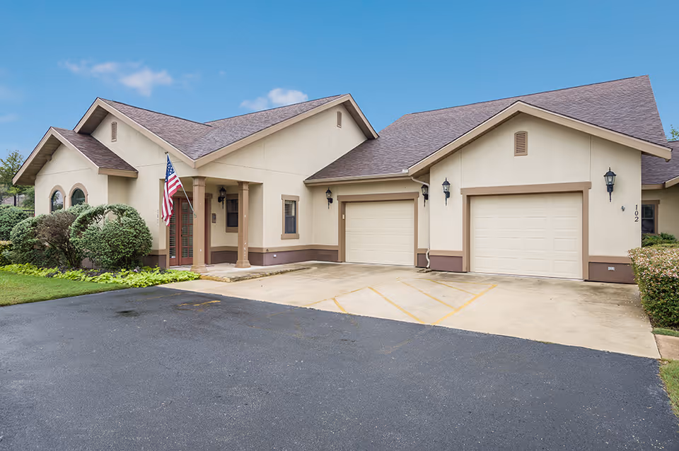 Exterior view of a single-story building with beige walls and brown trim, featuring two garage doors, an American flag near the entrance, and well-maintained landscaping with bushes and grass under a partly cloudy sky.