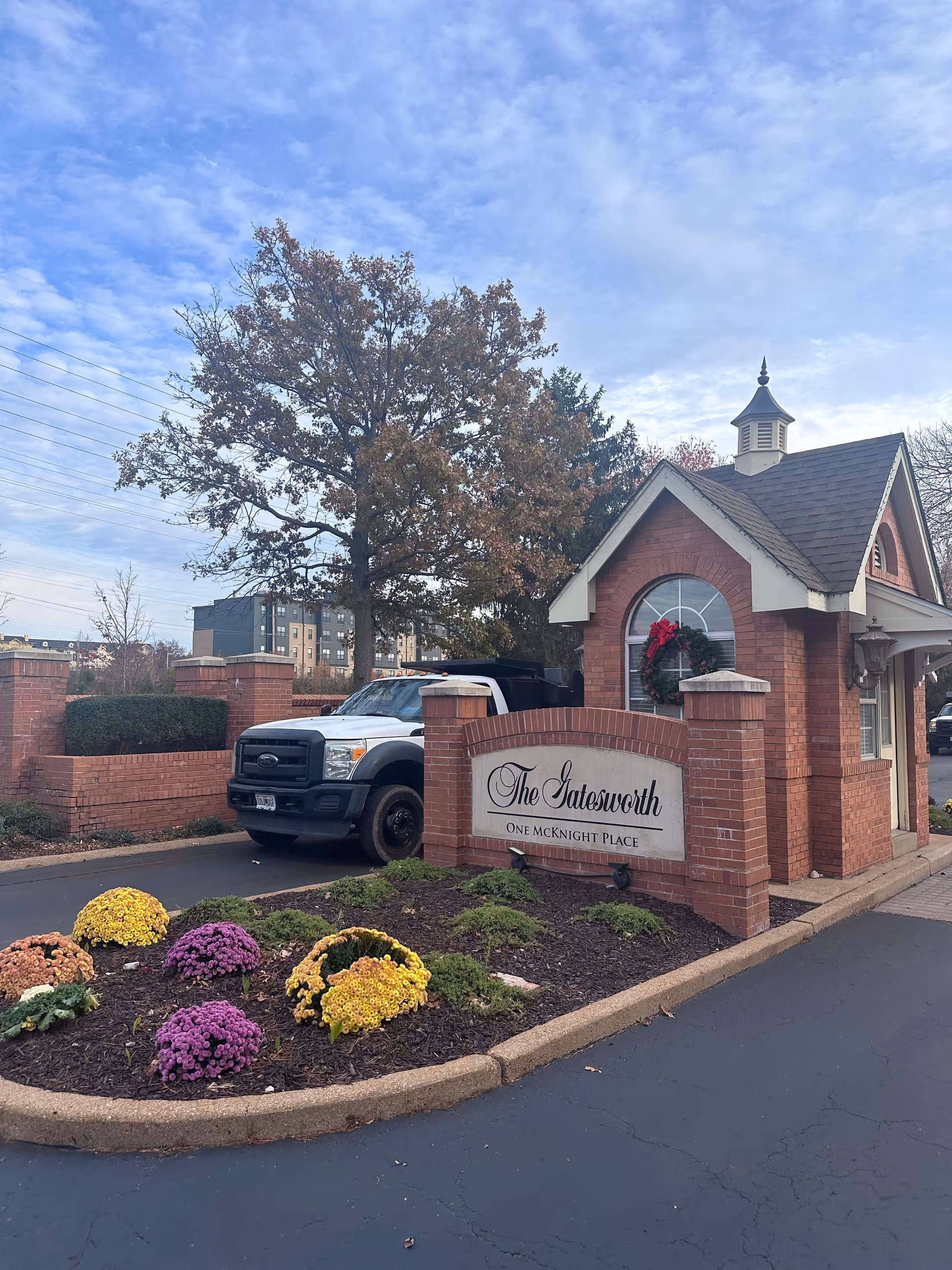 Entrance gatehouse of The Gatesworth facility with a brick structure, a sign reading 'The Gatesworth One McKnight Place', a wreath on the window, a white truck parked behind the gatehouse, and a landscaped flower bed with colorful flowers in front.