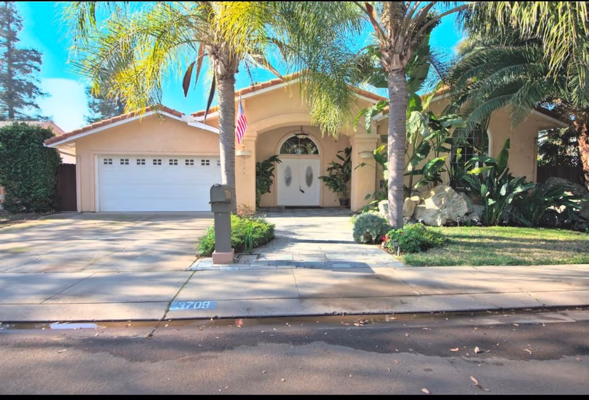 Front exterior view of a single-story house with a two-car garage, a driveway, palm trees, and various plants and shrubs in the front yard under a clear blue sky.