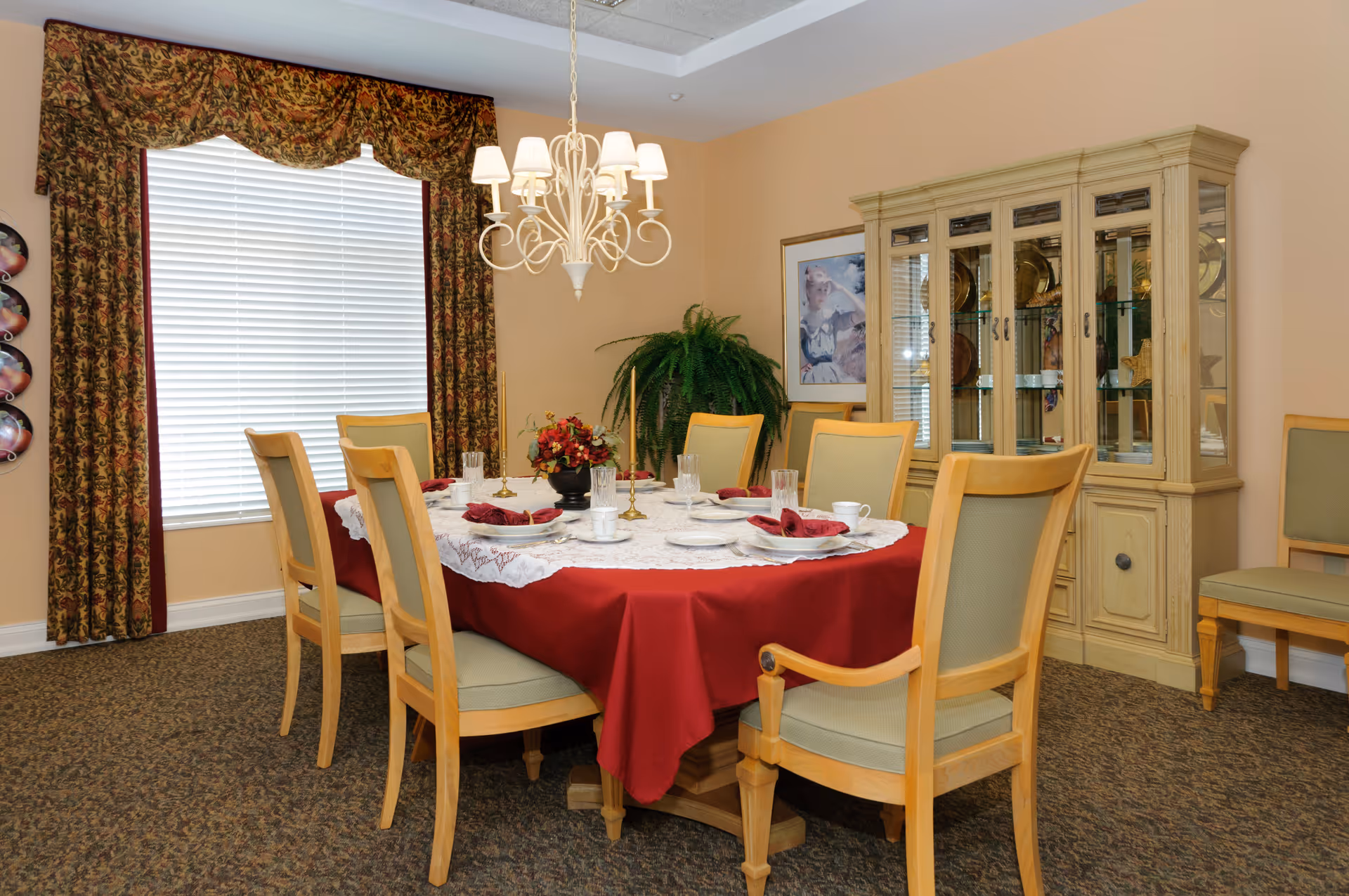 A dining room with a round table covered with a red tablecloth and a white lace overlay. The table is set with plates, glasses, cups, and red napkins. Eight wooden chairs with light green cushions surround the table. A chandelier with multiple lights hangs above the table. There is a large window with floral curtains and a valance, a wooden china cabinet with glass doors displaying plates, a framed picture on the wall, and a green potted plant in the corner.