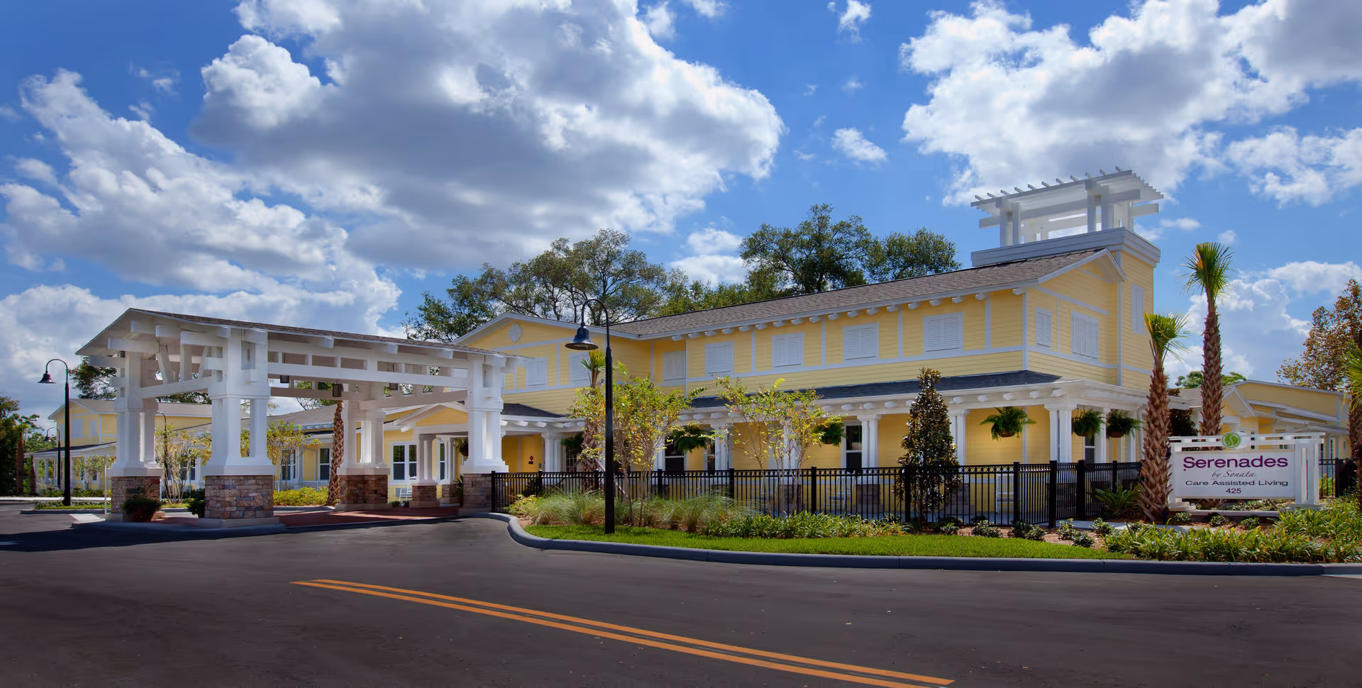 Exterior view of Serenades at Longwood, a yellow two-story assisted living facility with white trim, a covered entrance, landscaped greenery, palm trees, and a partly cloudy blue sky.