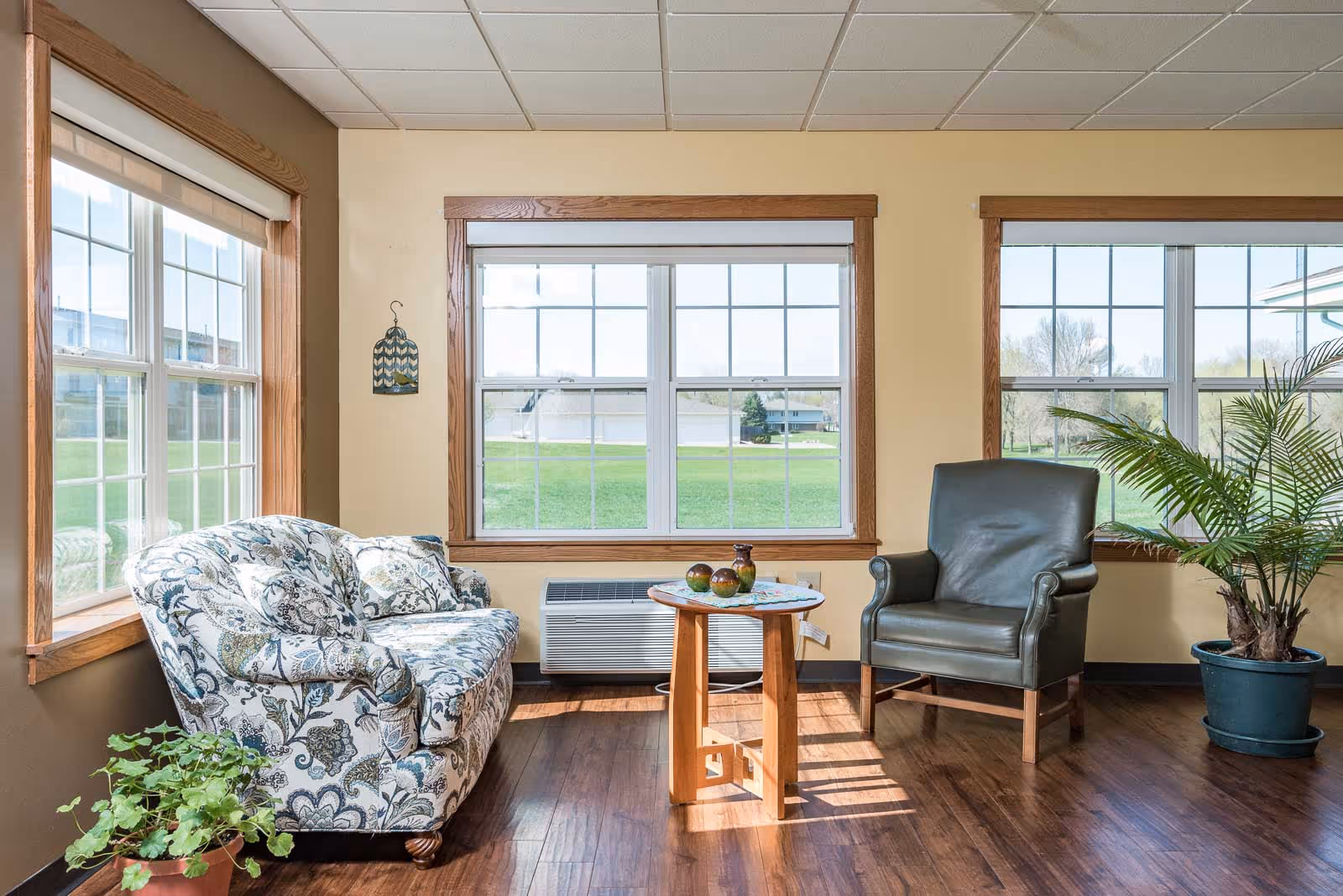 A bright sitting area with large windows showing a green lawn outside. The room has a floral patterned sofa on the left, a dark leather armchair on the right, a small wooden table with decorative items in the center, and two potted plants on the floor. The walls are beige with wooden trim around the windows.
