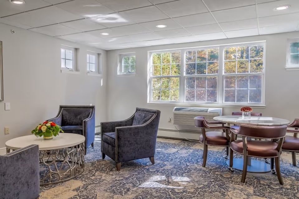 A bright and cozy sitting area in a senior living facility with large windows letting in natural light. The room features three dark upholstered armchairs around a round white coffee table with a floral arrangement, and a round dining table with four brown leather chairs. The floor is covered with a patterned carpet, and there is an air conditioning unit under the windows.