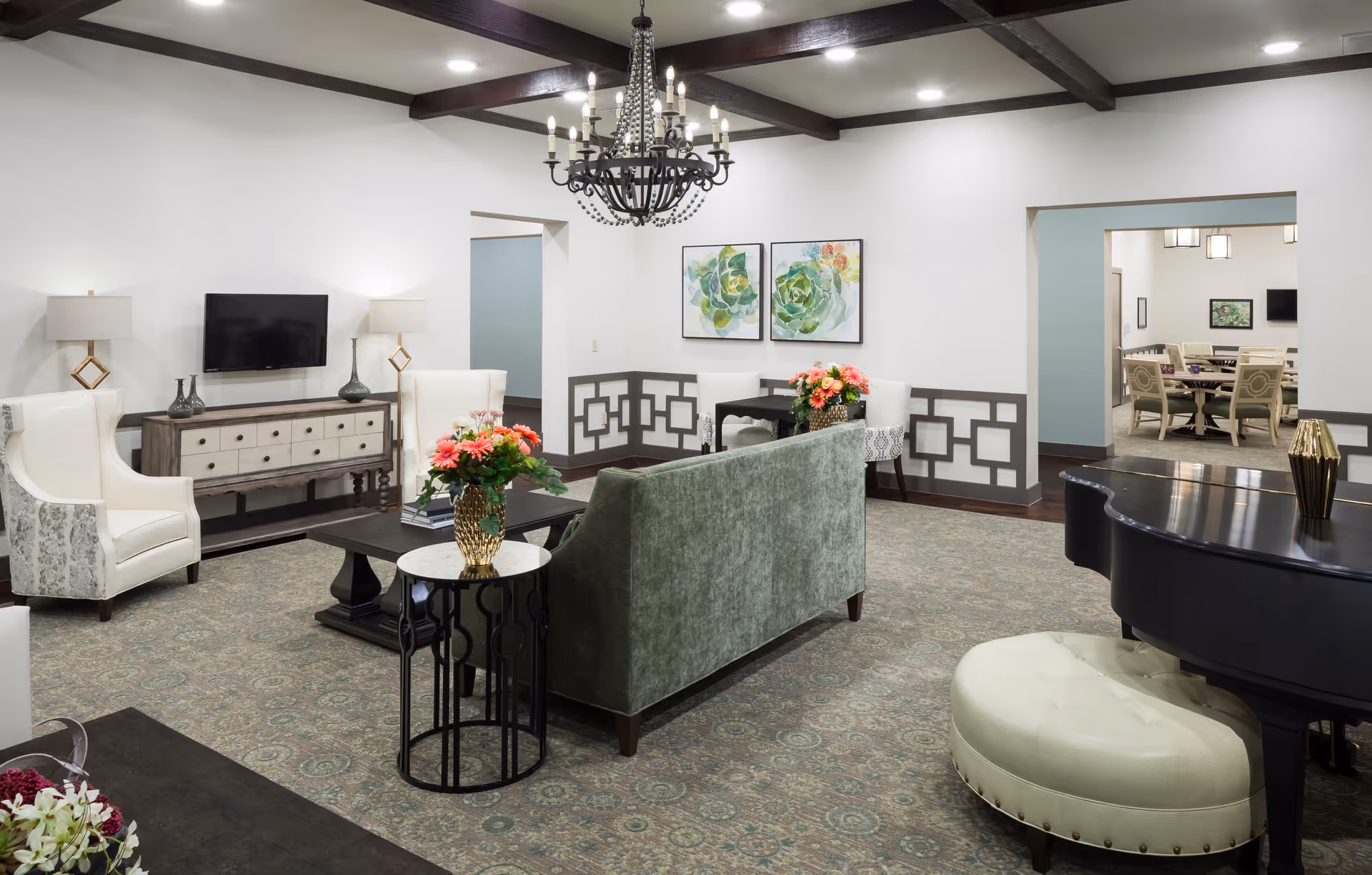 A spacious and elegantly decorated living room with a green upholstered sofa, two white armchairs, a dark wood coffee table, and a side table with a vase of flowers. The room features a patterned carpet, a chandelier hanging from a ceiling with dark wooden beams, and a wall-mounted flat-screen TV above a wooden console. In the background, there is a small dining area with two white chairs and a table, and another room with a larger dining table and chairs is visible through an open doorway. A black grand piano with a cream-colored cushioned bench is also present in the foreground.
