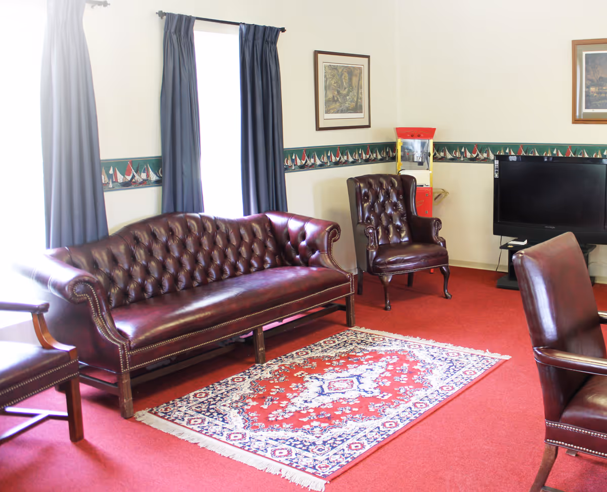 A living room with red carpet, a patterned area rug, a dark brown leather tufted sofa, two matching leather armchairs, a flat-screen TV on a stand, two framed pictures on the wall, and a popcorn machine in the corner. The room has white walls with a decorative border featuring sailboats and two windows with dark curtains.