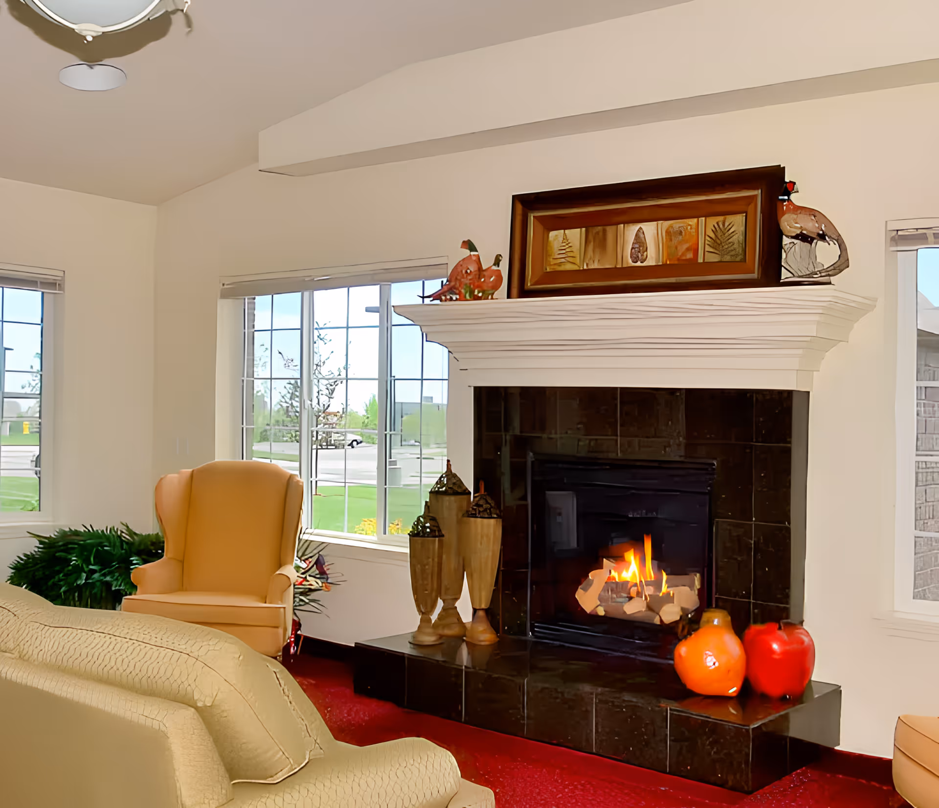 A cozy living room with a lit fireplace and mantel decorations, upholstered armchairs, and large windows letting in daylight.