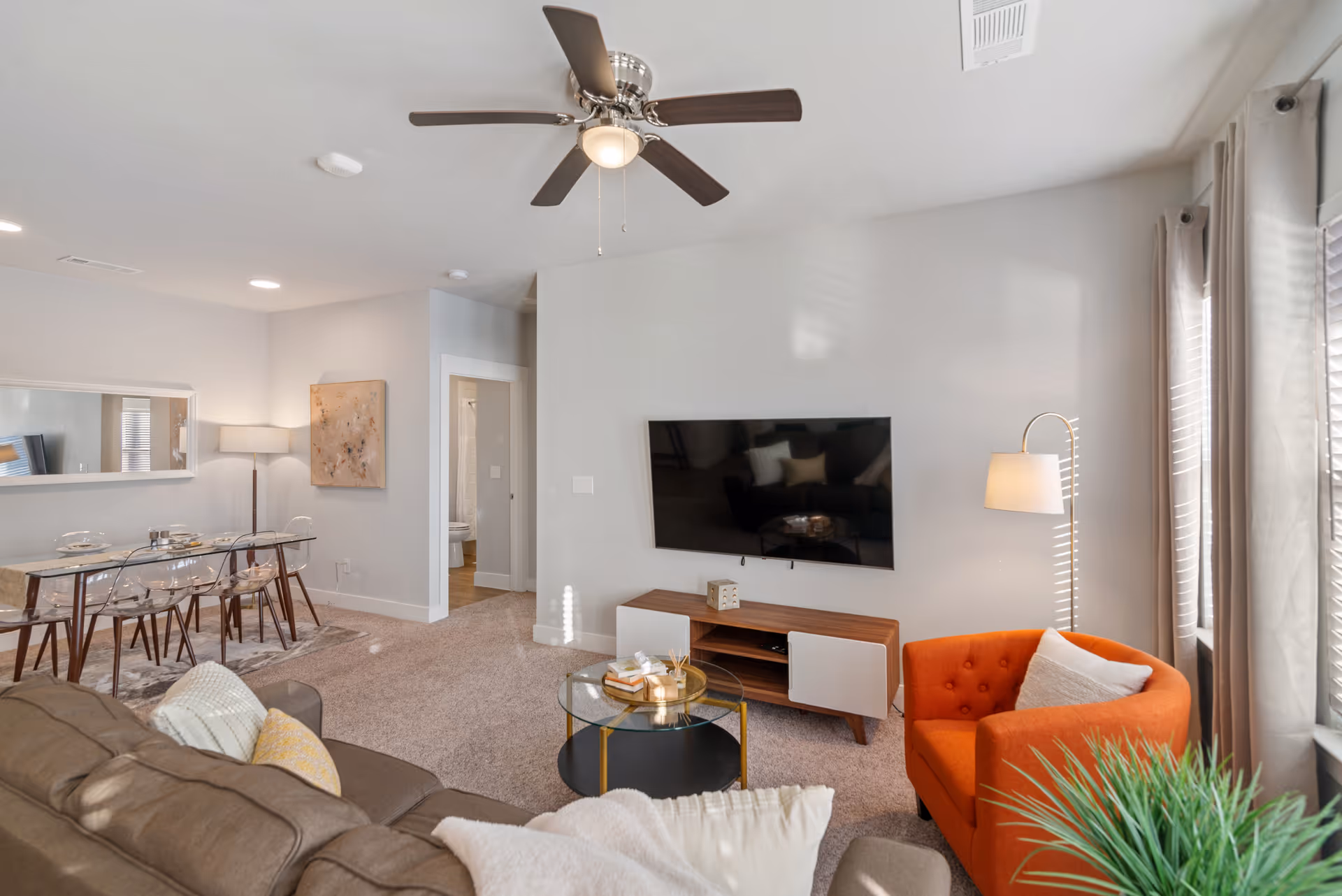Bright living room with a wall-mounted TV, brown sofa, orange armchair, glass coffee table and adjacent dining area.
