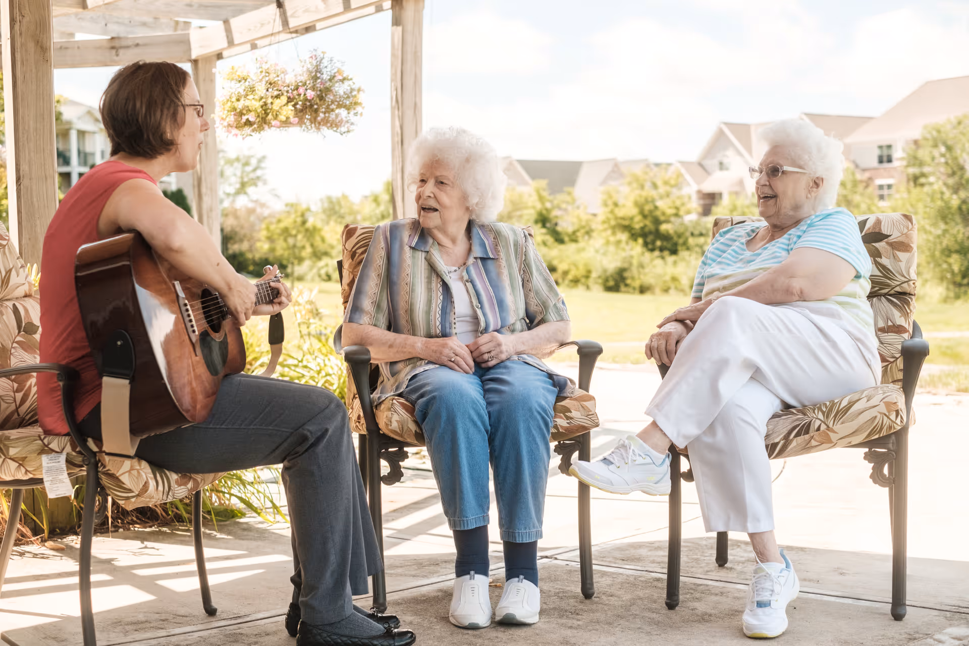 An outdoor patio scene at a senior living facility where two elderly women are seated on cushioned chairs, engaged in conversation with a woman playing an acoustic guitar. The setting is bright and sunny with greenery and residential buildings in the background.