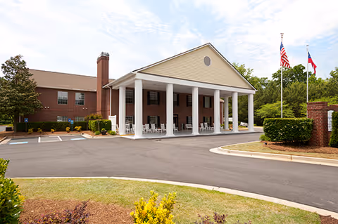 Front exterior of a senior living building with a large covered entrance supported by white columns, flagpoles, and a circular driveway.