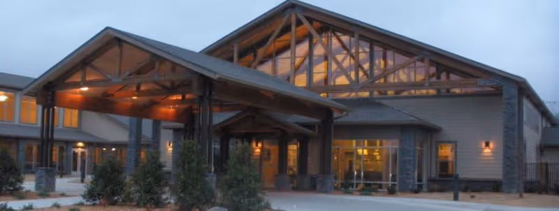 Exterior view of RiverLodge Assisted Living facility at dusk, showing the main entrance with a large covered drop-off area supported by wooden beams and stone pillars, warm interior lights visible through windows.