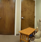Interior view showing two closed wooden doors with silver handles, a small wooden side table, and part of a patterned upholstered chair in a room with beige walls and carpeted floor.