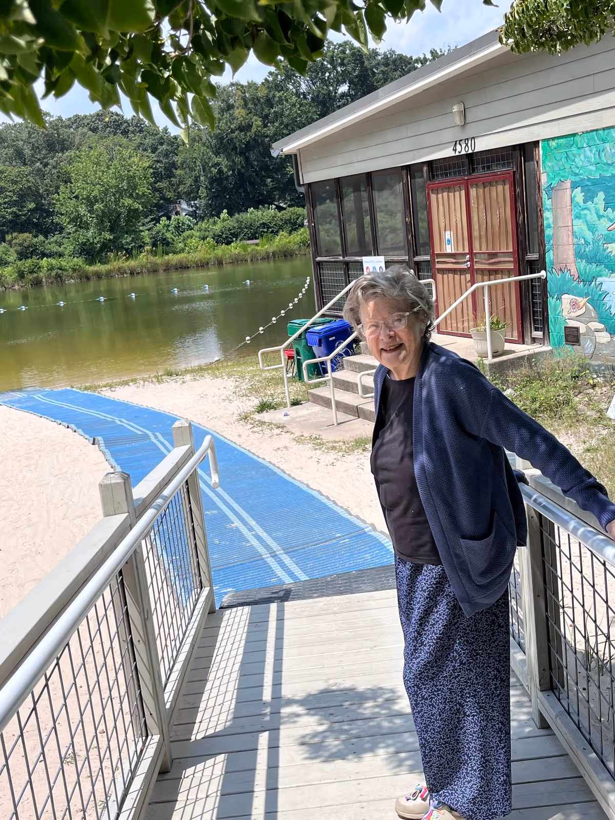 An elderly woman standing on a wooden ramp with metal railings near a sandy beach area. Behind her is a building with large windows and a mural painted on the wall. In the background, there is a calm body of water surrounded by trees and greenery.