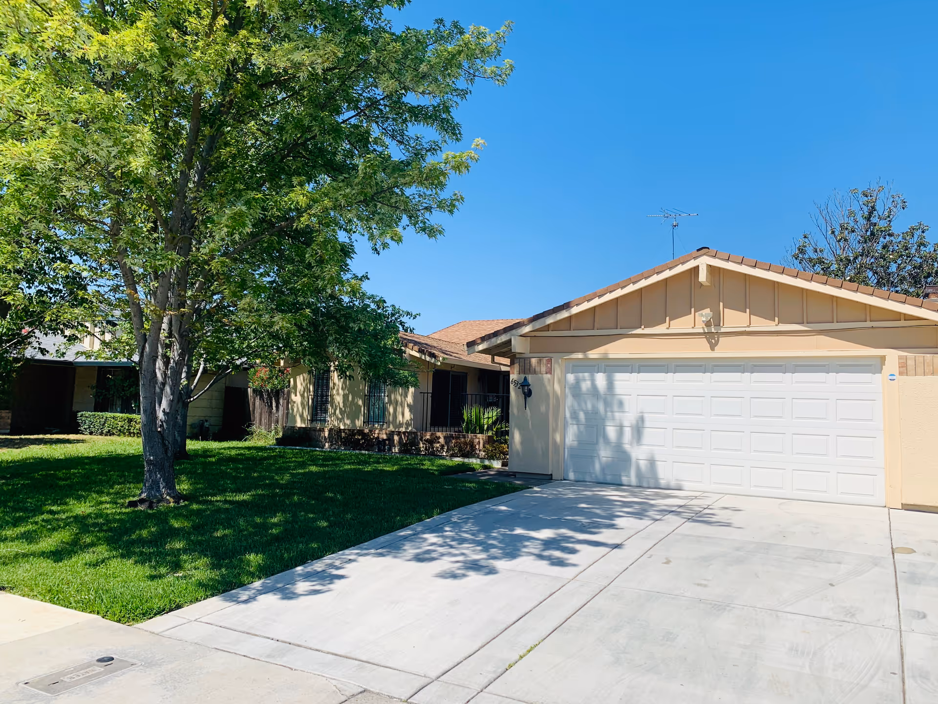 A single-story house with a beige exterior and a white garage door. There is a large green tree and well-maintained grass in the front yard under a clear blue sky.