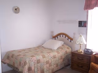 A small bedroom with a single bed covered by a patchwork quilt. There is a white pillow on the bed. Next to the bed is a wooden nightstand with a lamp and a small radio. A window with pink curtains is partially visible on the right side of the image. The walls are plain white with minimal decoration.