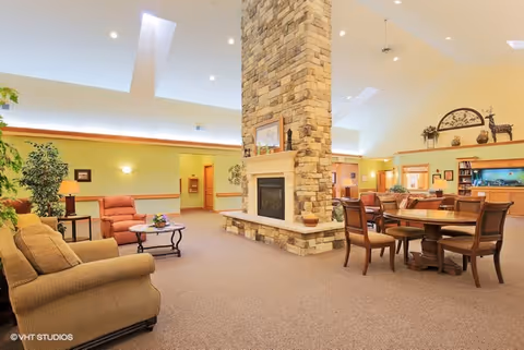 Spacious senior living common area with a tall stone fireplace in the center, surrounded by comfortable seating including sofas and armchairs, a round dining table with chairs, and decorative plants and artwork on the walls.
