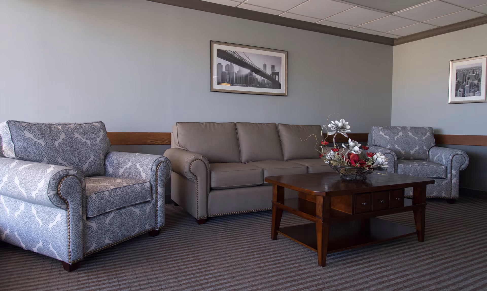 A living room area with a gray sofa and two patterned armchairs arranged around a wooden coffee table with a floral centerpiece. The walls are painted gray and decorated with two framed black and white cityscape photographs.