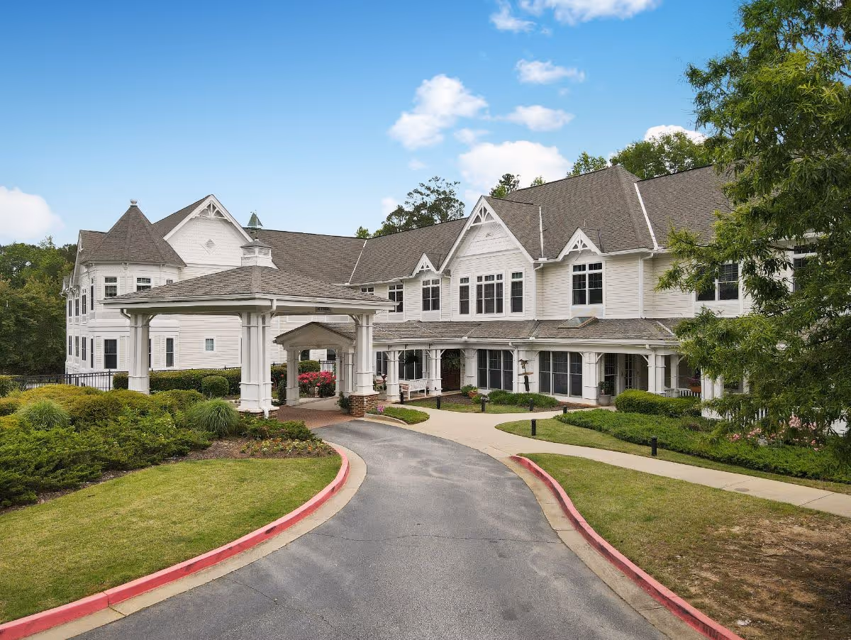 Front exterior view of a large, white assisted living facility building with a covered entrance driveway, surrounded by well-maintained landscaping and greenery under a blue sky with some clouds.