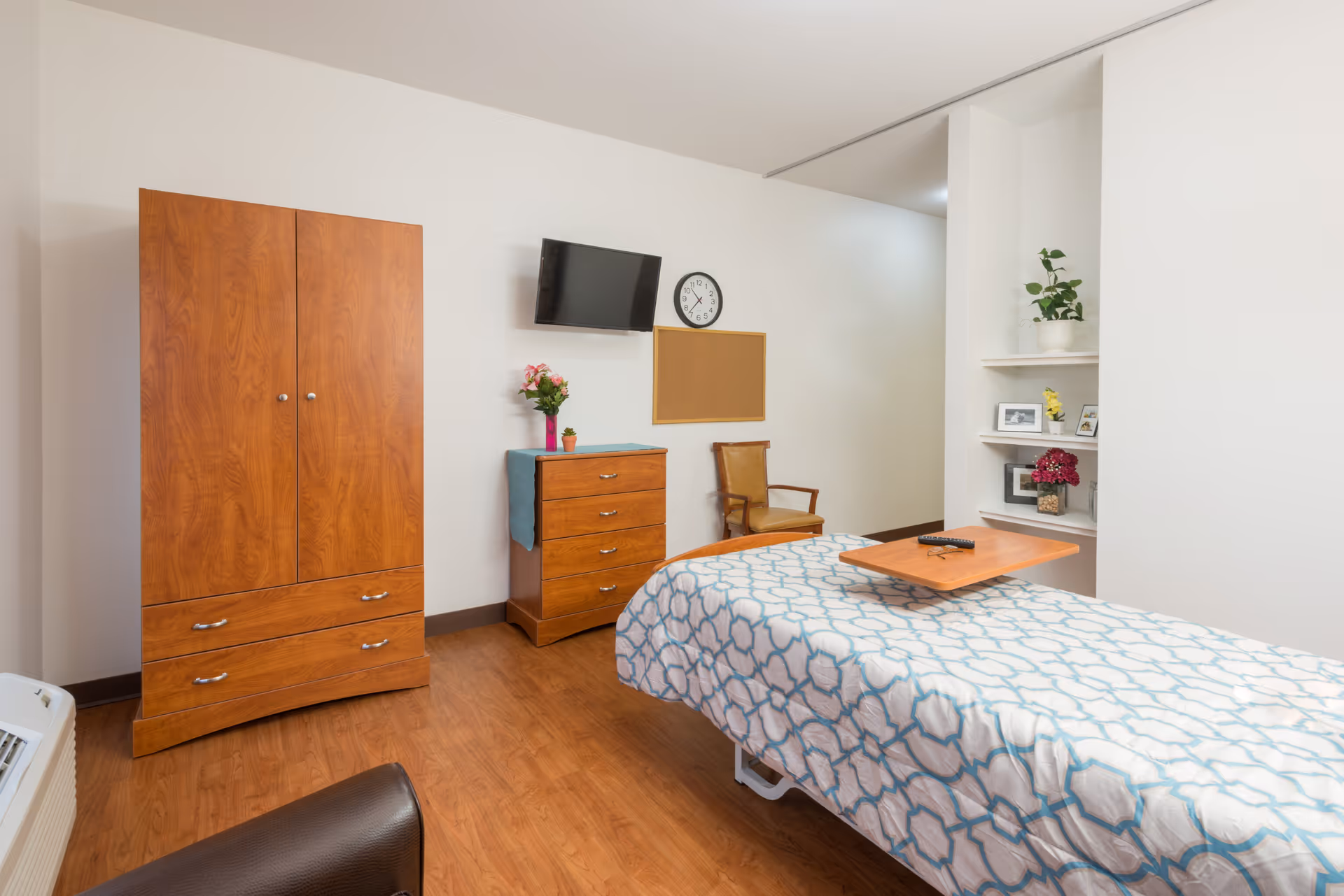 A simple furnished patient bedroom with a bed, wooden wardrobe and dresser, wall-mounted TV, chair, and decorative shelves.