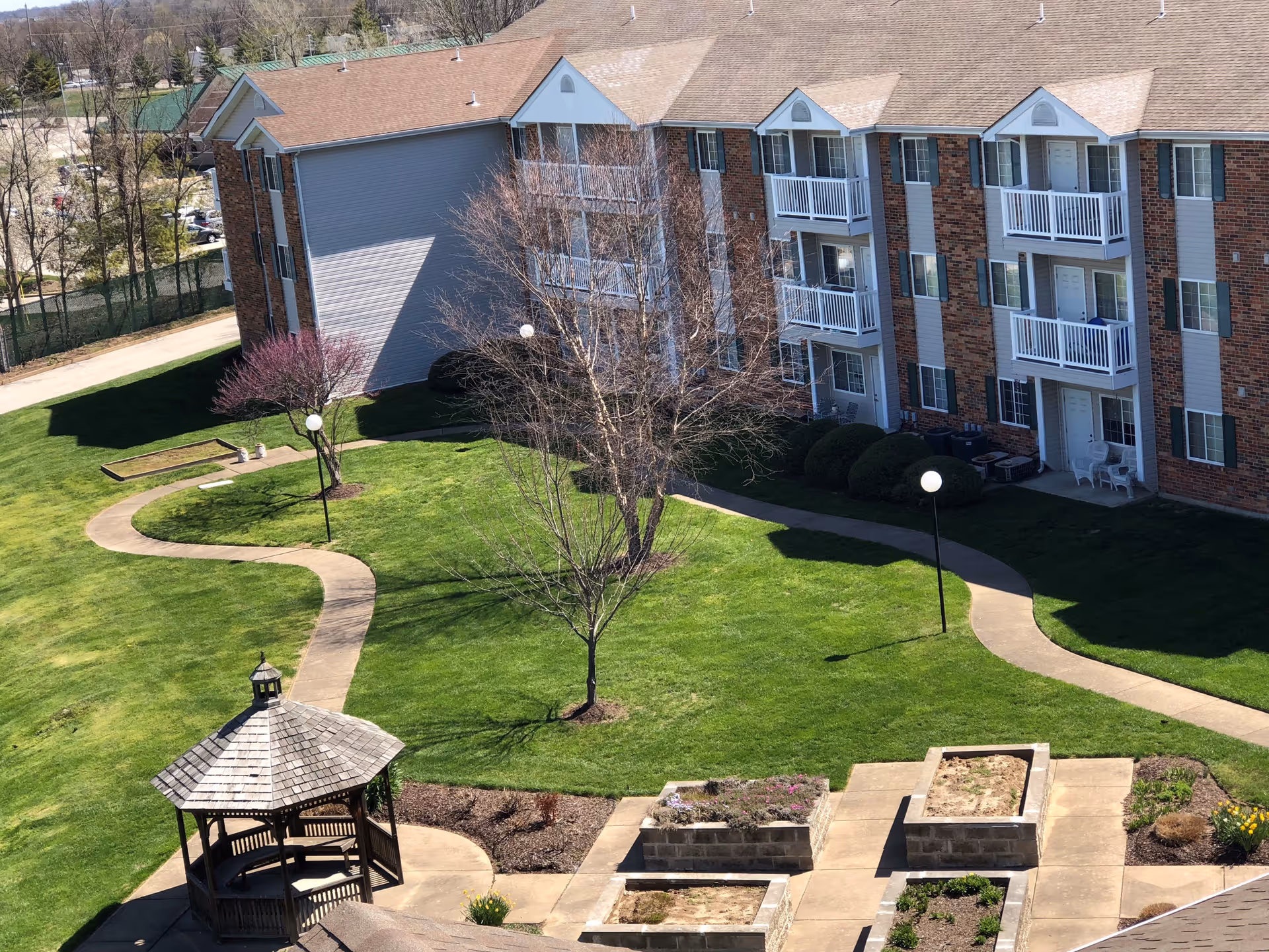 Outdoor view of a senior living facility with a well-maintained lawn, a wooden gazebo, paved walking paths, several trees, and a three-story brick building with balconies and white railings.