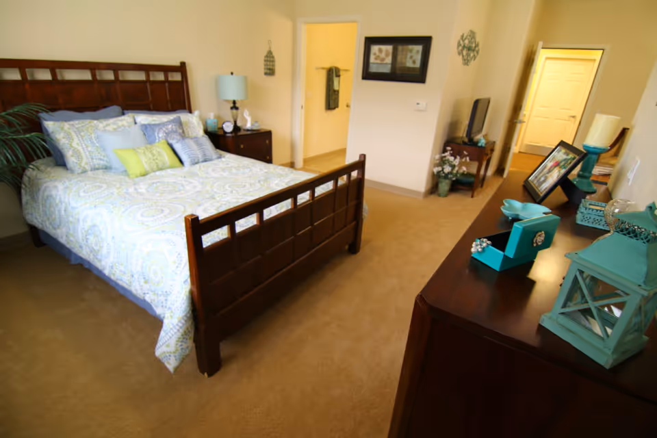 A cozy bedroom with a wooden bed frame and a bed made with patterned bedding and multiple pillows. There is a wooden nightstand with a lamp and decorative items next to the bed. A dresser with various decorative objects, including a turquoise lantern and a jewelry box, is visible in the foreground. The room has beige carpet and walls, with two doorways leading to other areas.