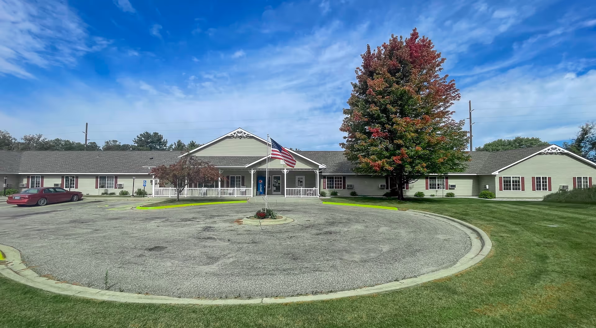 Front exterior view of a single-story senior living facility building with a circular driveway, an American flag on a flagpole in the center, a large tree with autumn-colored leaves to the right, and a red car parked on the left side. The building has light green siding, white trim, and red shutters on the windows under a partly cloudy blue sky.