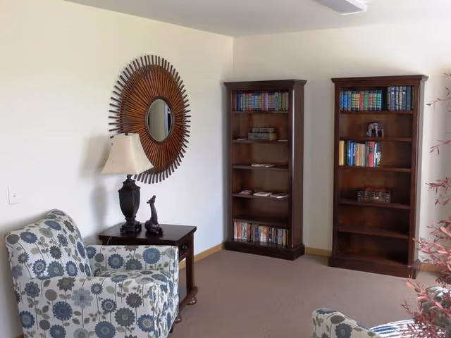 A cozy living room area with two floral patterned armchairs, a wooden side table with a lamp and decorative figurine, a round sunburst mirror on the wall, and two tall wooden bookshelves filled with books and decorative items.