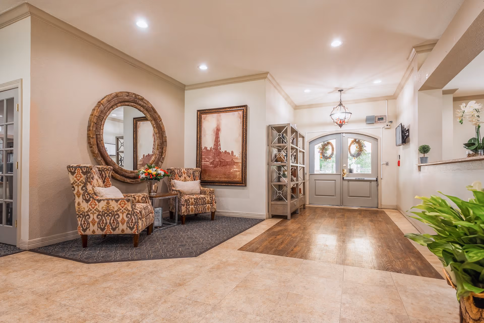 A bright and welcoming senior living facility lobby with two patterned armchairs and a small table with flowers on the left, a large round wooden-framed mirror above the chairs, a framed artwork on the wall, a metal shelving unit, and double doors decorated with wreaths at the far end. The floor transitions from tile to wood, and there are plants and a reception counter on the right side.