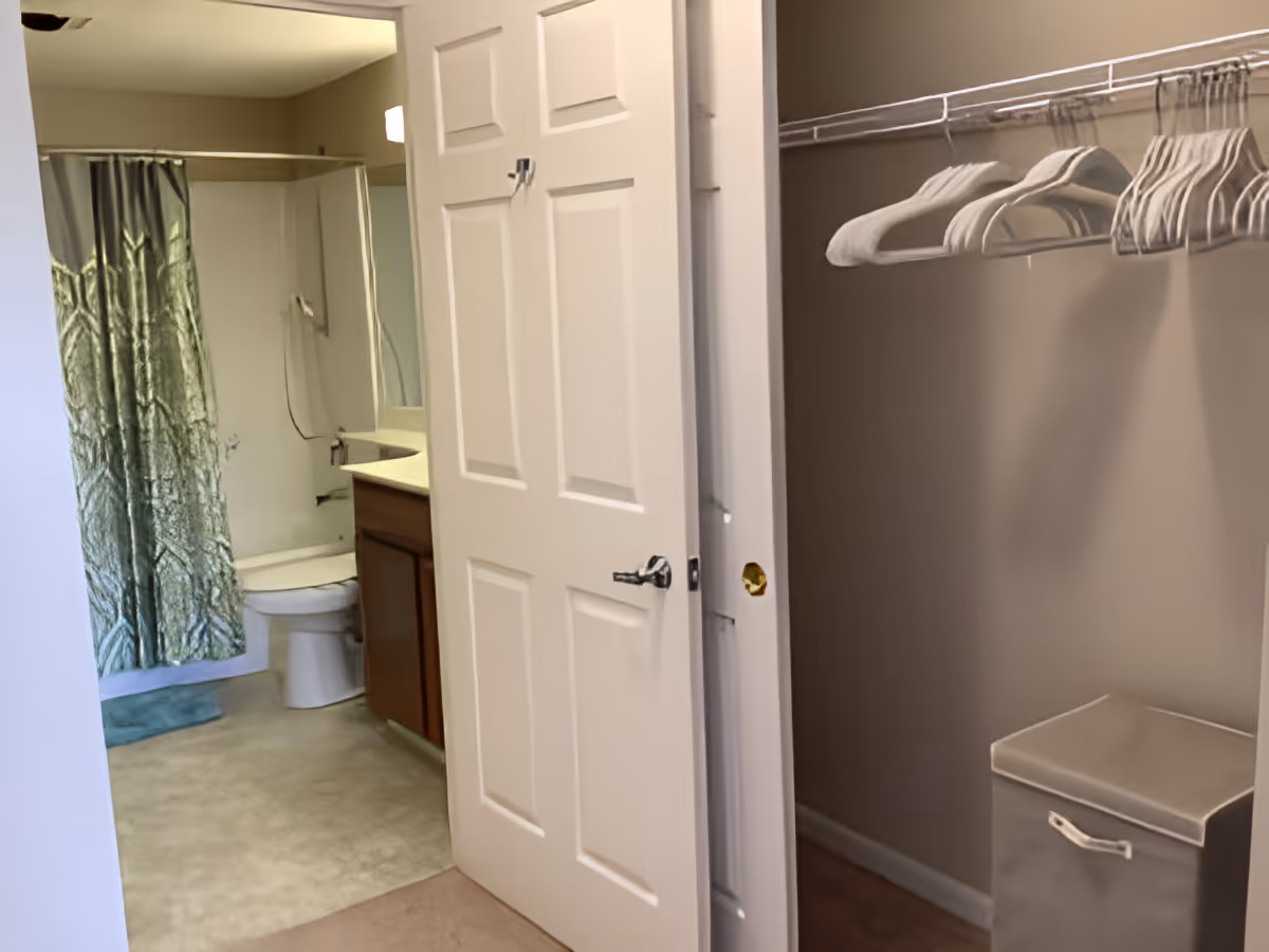 View of a bathroom with a shower curtain, toilet, and sink with a mirror, seen through an open door. To the right, there is a closet with a hanging rod holding several empty hangers and a laundry hamper on the floor.