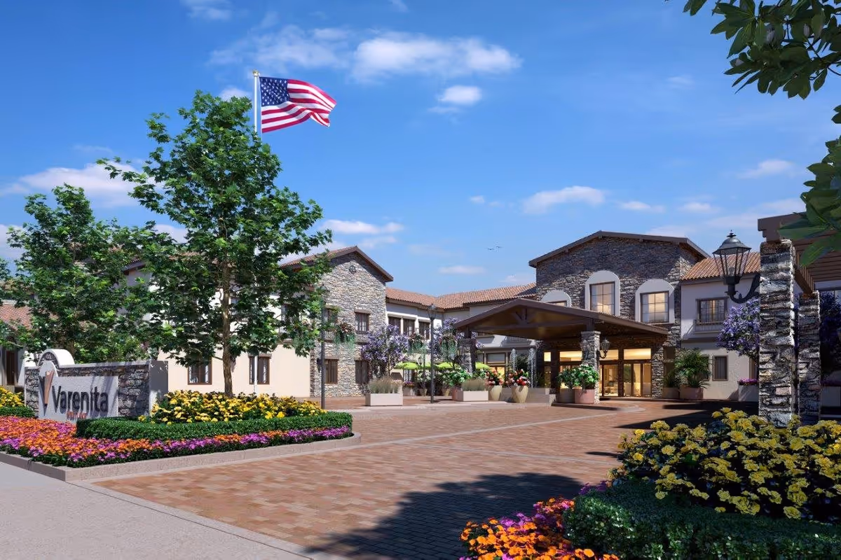 Exterior view of Varenita Westlake facility with a stone and stucco building, landscaped flower beds, trees, and an American flag flying on a flagpole under a blue sky with some clouds.