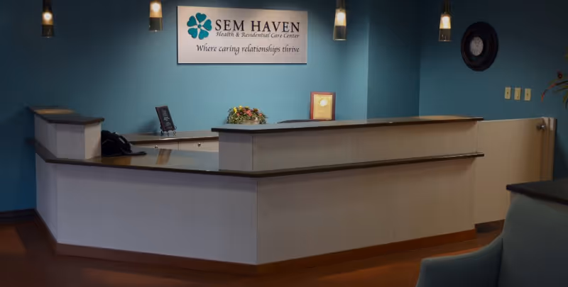 Reception desk area inside SEM Haven Health & Residential Care Center with a sign on the wall that reads 'SEM HAVEN Health & Residential Care Center Where caring relationships thrive'. The desk has a phone, a small framed sign, and a flower arrangement. The walls are painted blue and there is a clock on the wall.