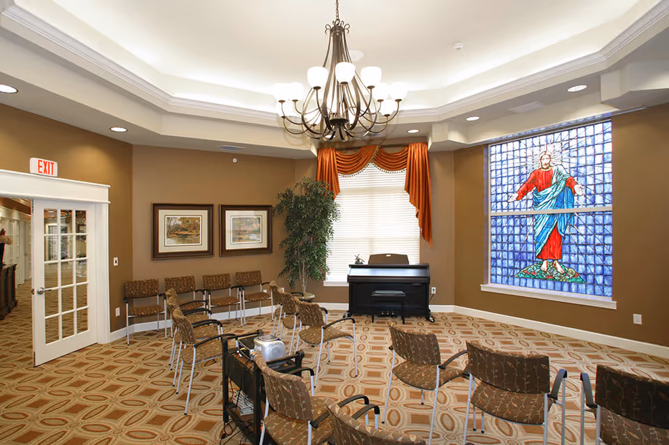 A room with a chandelier hanging from a tray ceiling, several chairs arranged in rows, a piano against the wall, a large stained glass window depicting a figure in red and blue robes, two framed pictures on the wall, a potted plant, and an exit door with glass panels.