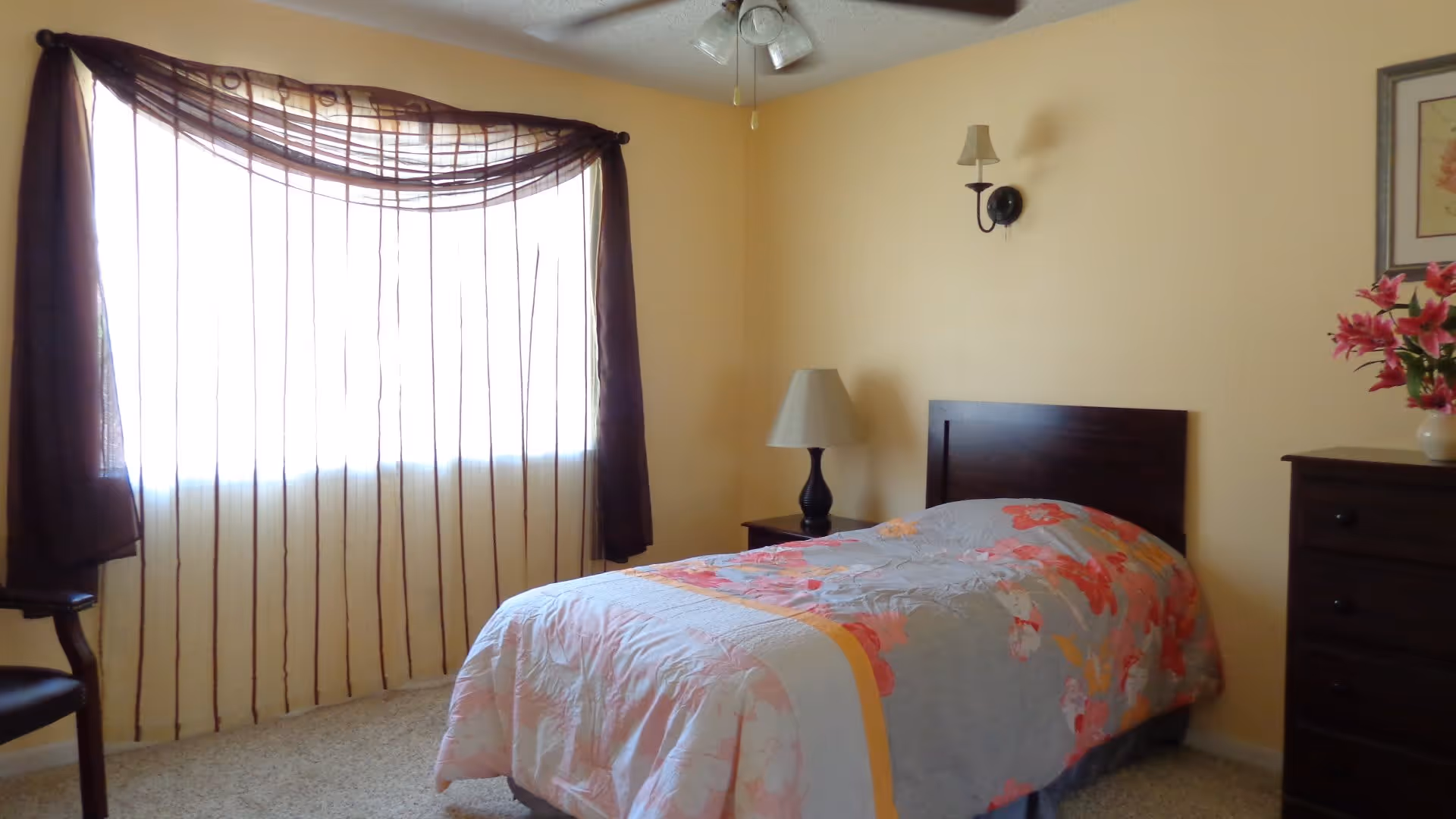 A bedroom with a single bed covered in a floral patterned comforter. There is a wooden nightstand with a lamp next to the bed, a wooden dresser with a vase of pink flowers, a chair near a window with sheer brown curtains, and a ceiling fan with lights.