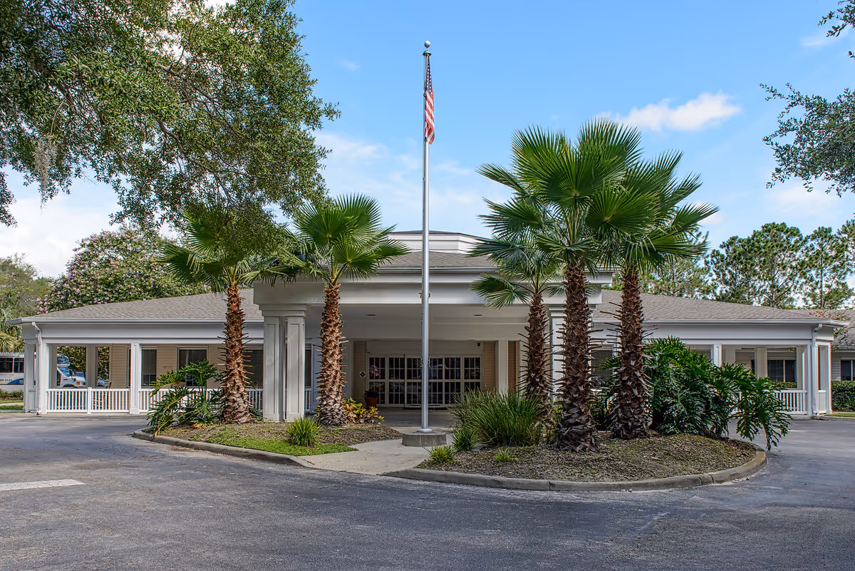 Front exterior view of a single-story senior living facility building with a covered entrance, surrounded by palm trees and other greenery, under a blue sky with some clouds. An American flag is flying on a flagpole in front of the entrance.
