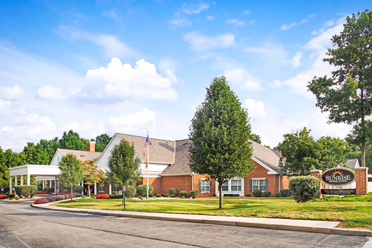 Exterior view of a senior living facility building with a brick facade, surrounded by green trees and well-maintained landscaping under a partly cloudy blue sky. There is a sign in front that reads 'Sunrise Senior Living'.