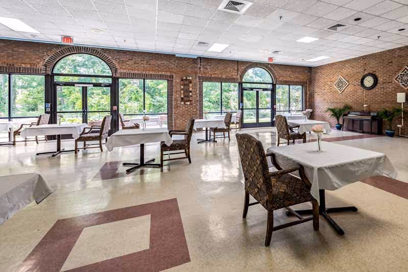 A spacious dining room with multiple tables covered in white tablecloths, each accompanied by cushioned chairs. Large arched windows and glass doors allow natural light to fill the room, revealing a green outdoor view. The walls are brick, and there is a piano and some decorative plants in the corner.