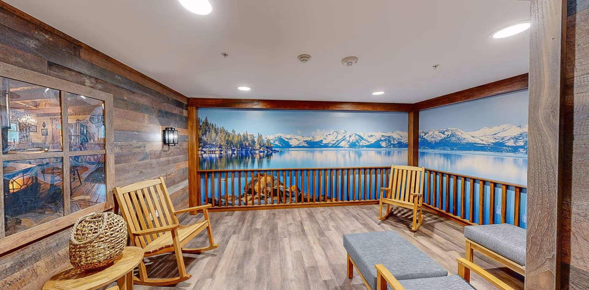 A cozy indoor seating area with wooden rocking chairs and cushioned benches. The walls feature wood paneling and a large mural of a serene lake with snow-capped mountains and pine trees, creating a calming atmosphere.