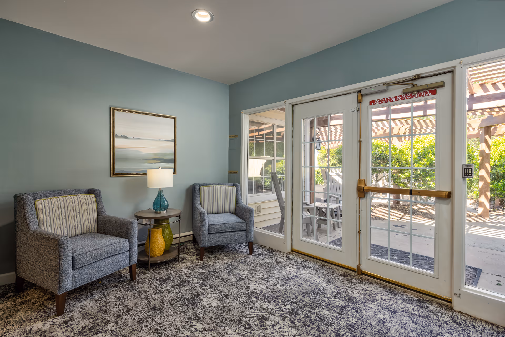 Small seating area with two upholstered armchairs, a side table and lamp beside glass doors leading to a sunlit outdoor patio.