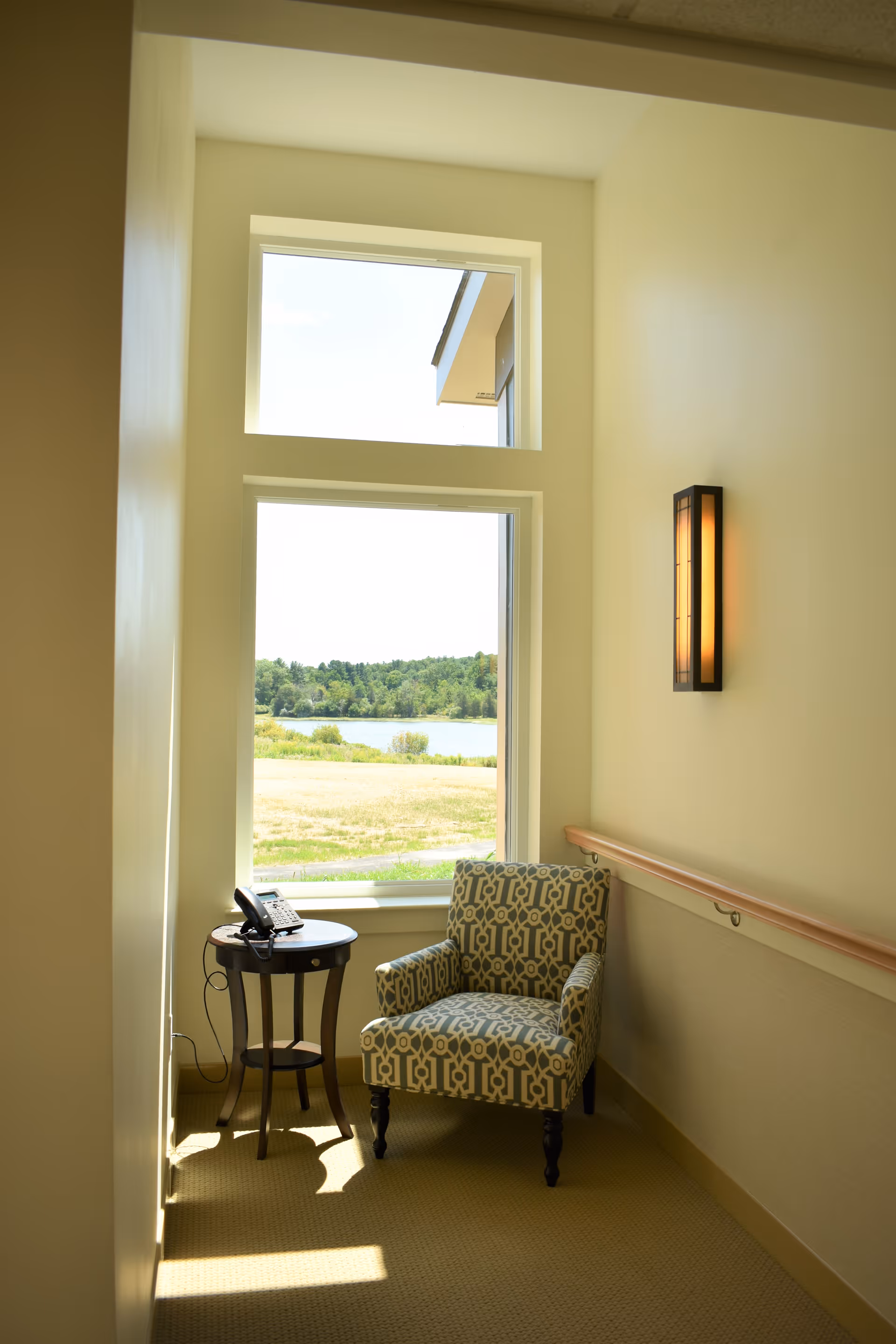 A cozy corner with a patterned armchair and a small round wooden table holding a telephone, positioned next to a large window overlooking a grassy area and a body of water with trees in the background. A modern wall sconce provides warm lighting on the right wall.