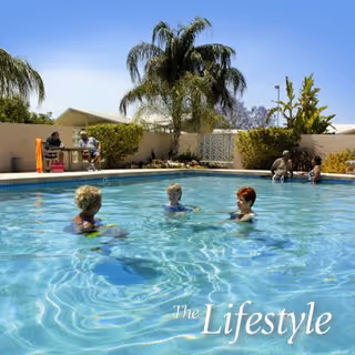 Several elderly people enjoying a sunny day in an outdoor swimming pool surrounded by palm trees and a beige wall, with some people sitting and relaxing poolside.